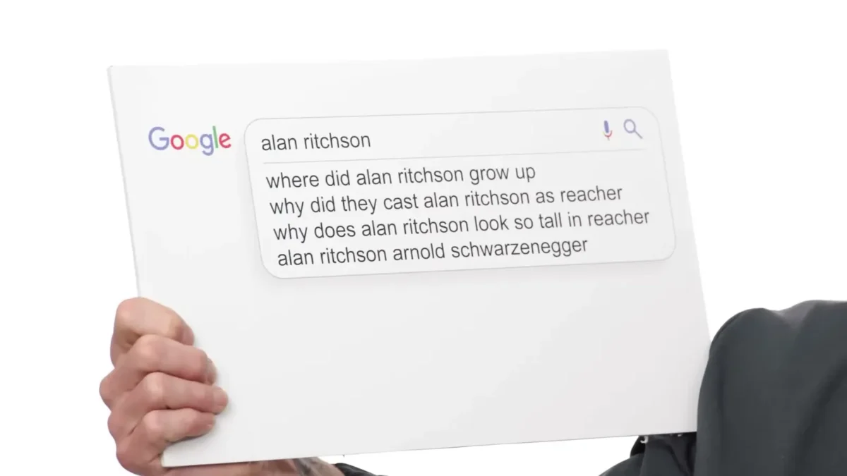 Hands holding a card showing Google search suggestions for Alan Ritchson, including why did they cast alan ritchson as reacher