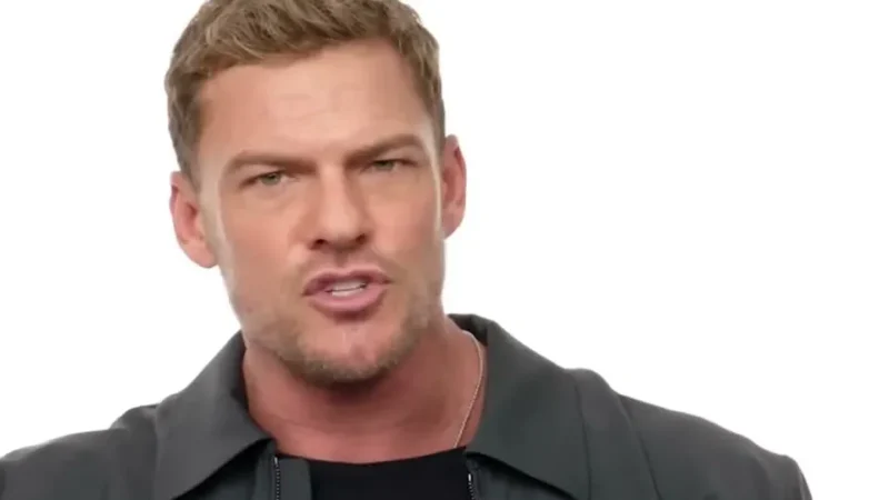 Alan Ritchson in a dark jacket and black T-shirt, gesturing as he talks to the camera against a white background