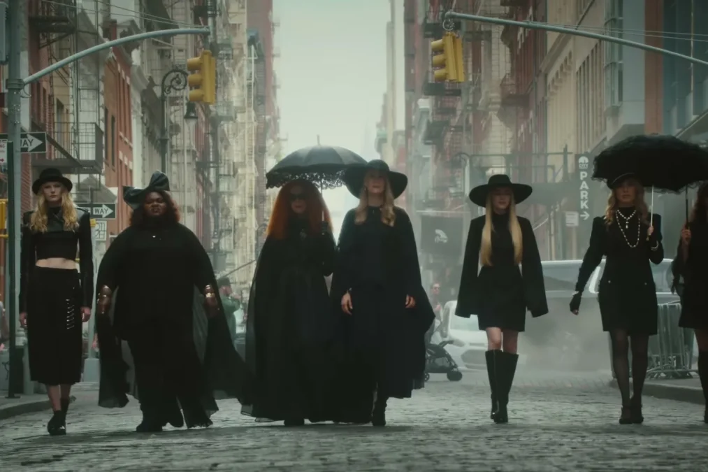 Seven women in black hats and coats, two with black umbrellas, walking down a cobblestone street in Manhattan in the American Horror Story 13 trailer