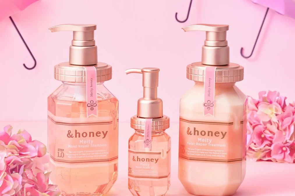 The pink &honey Melty Moist Repair shampoo, oil and treatment bottles against a pink background with pink umbrellas and flowers
