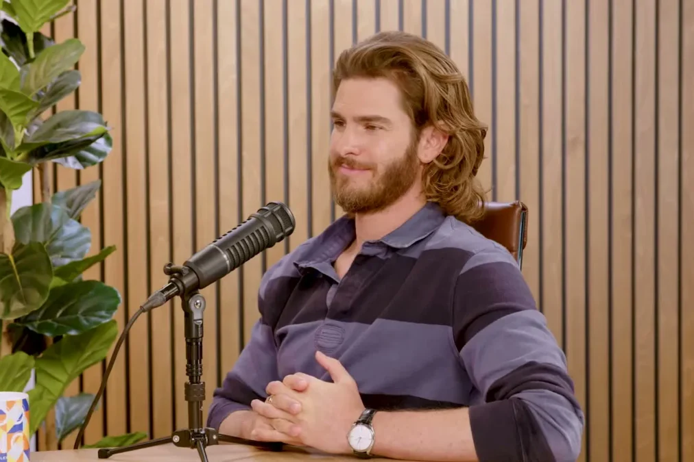 Andrew Garfield with long hair and a beard, in a striped rugby shirt, smiling beside a podcast microphone