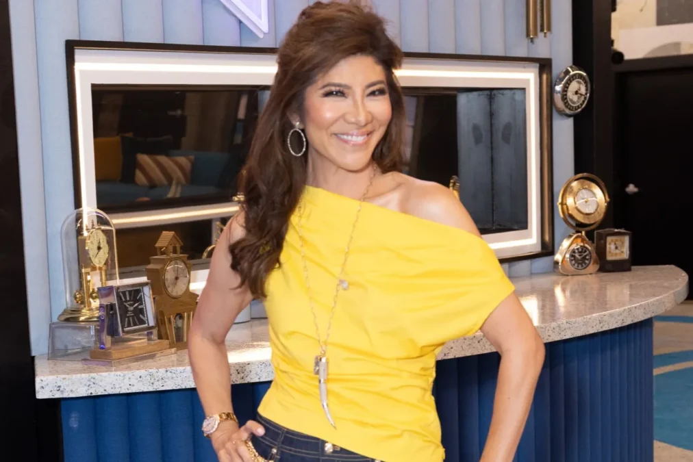 Julie Chen Moonves in a yellow top standing in the Big Brother 28 house beside a counter lined with clocks