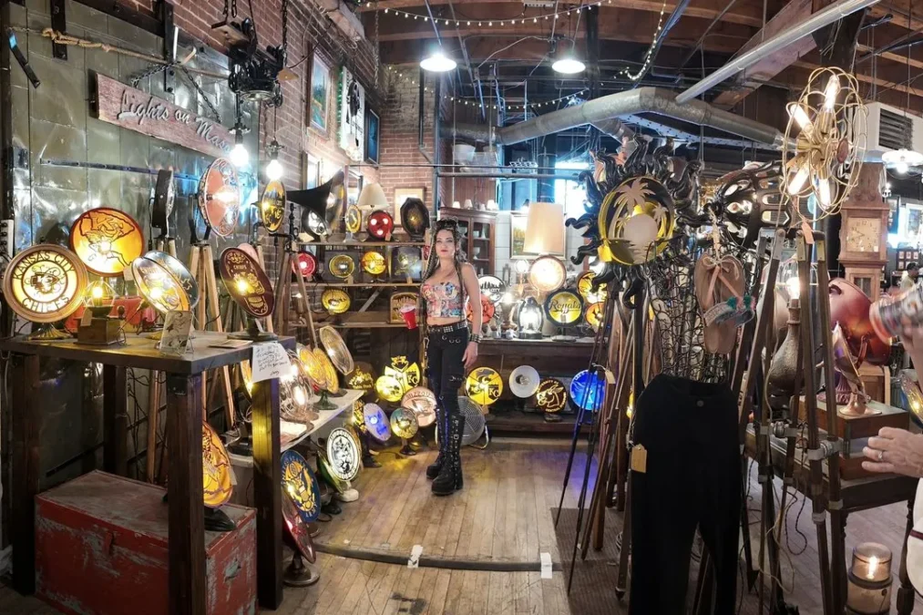 A vintage booth inside a West Bottoms warehouse, crowded with lit-up sports signs, old lamps and antiques under string lights and exposed beams, with shoppers browsing