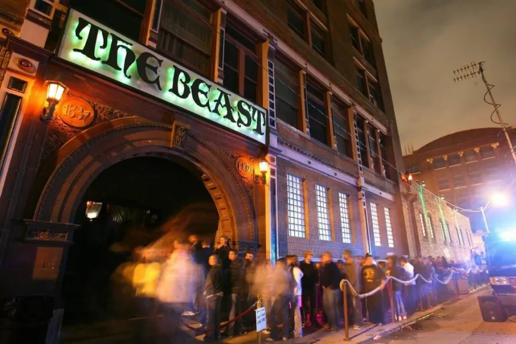 The Beast haunted house at night, its green neon sign over an arched brick entrance, with a long line of visitors along a rope outside