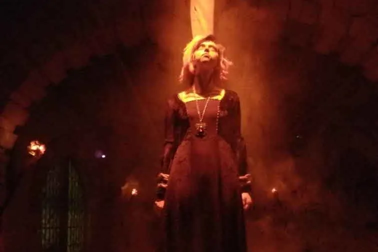 A woman in a dark period dress stands in orange fog on a lit platform in a witch-trial scene at the Beast