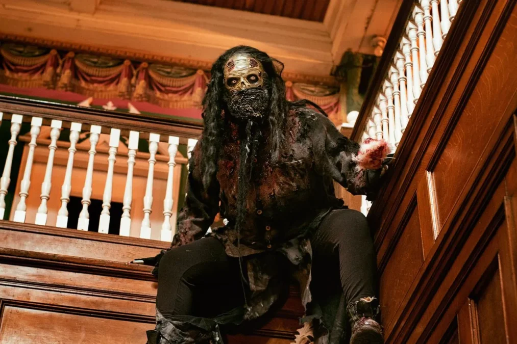 A masked, long-haired creature crouches on the wooden staircase of the Beast's ornate entrance hall