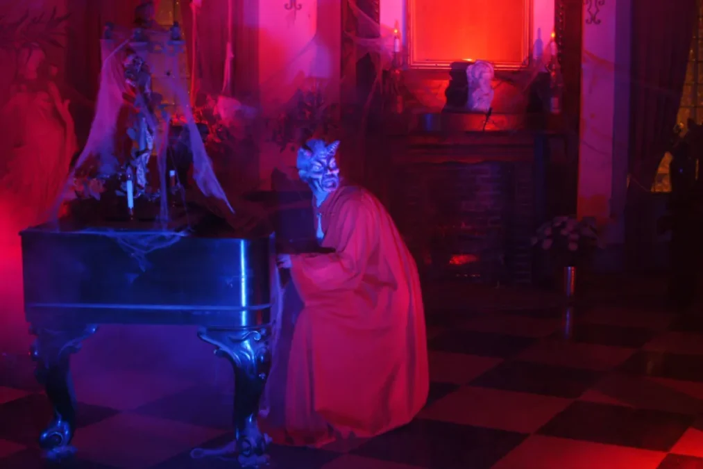 A red-lit haunted parlor with a cobwebbed piano, a checkered floor and a blue-faced figure in a red robe