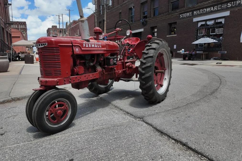 A red vintage Farmall tractor parked on a West Bottoms street in front of brick warehouses and the Stowe Hardware building on a sunny day