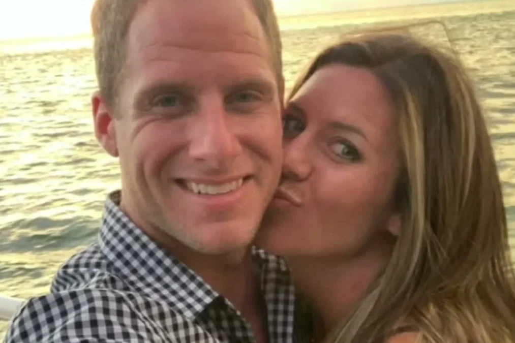 Caleb Flynn smiling as his wife, Ashley Flynn, kisses his cheek in a selfie by the water