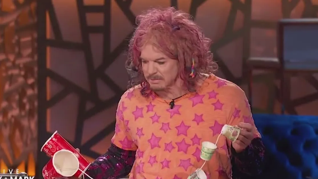 Carrot Top holding handmade props, a red cup and a small device, on a talk show couch