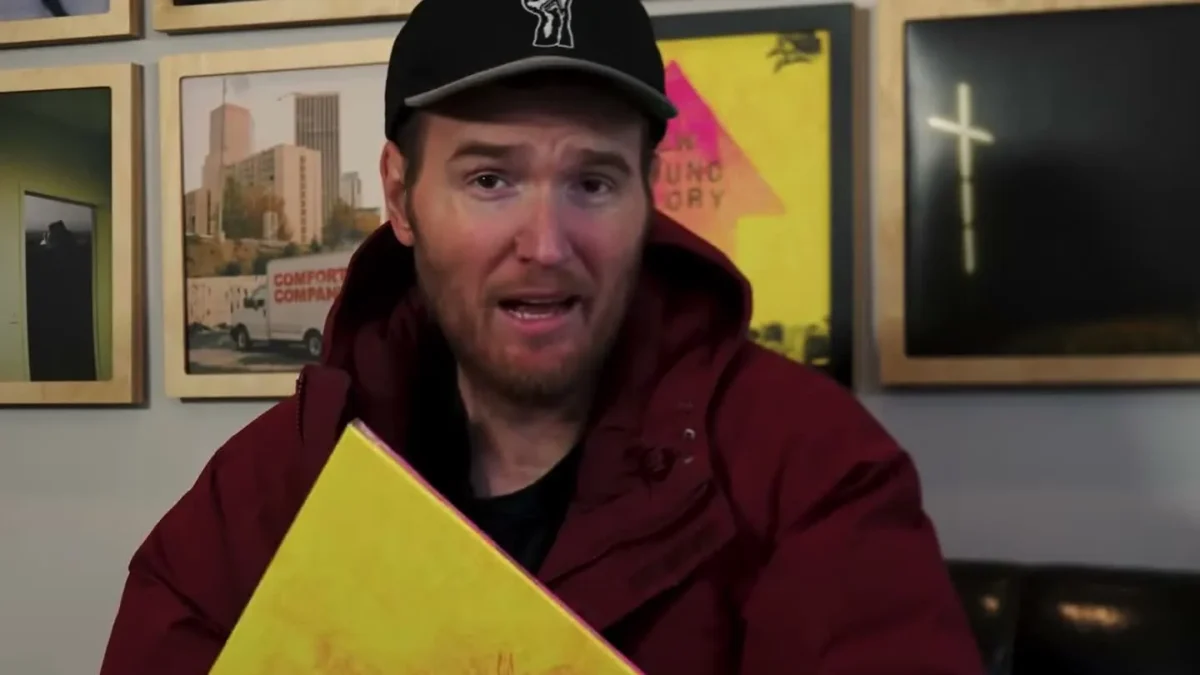 Chad Gilbert of New Found Glory, in a black cap and a maroon jacket, holding the band's album Listen Up!