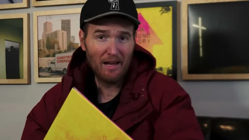 Chad Gilbert of New Found Glory, in a black cap and a maroon jacket, holding the band's album Listen Up!