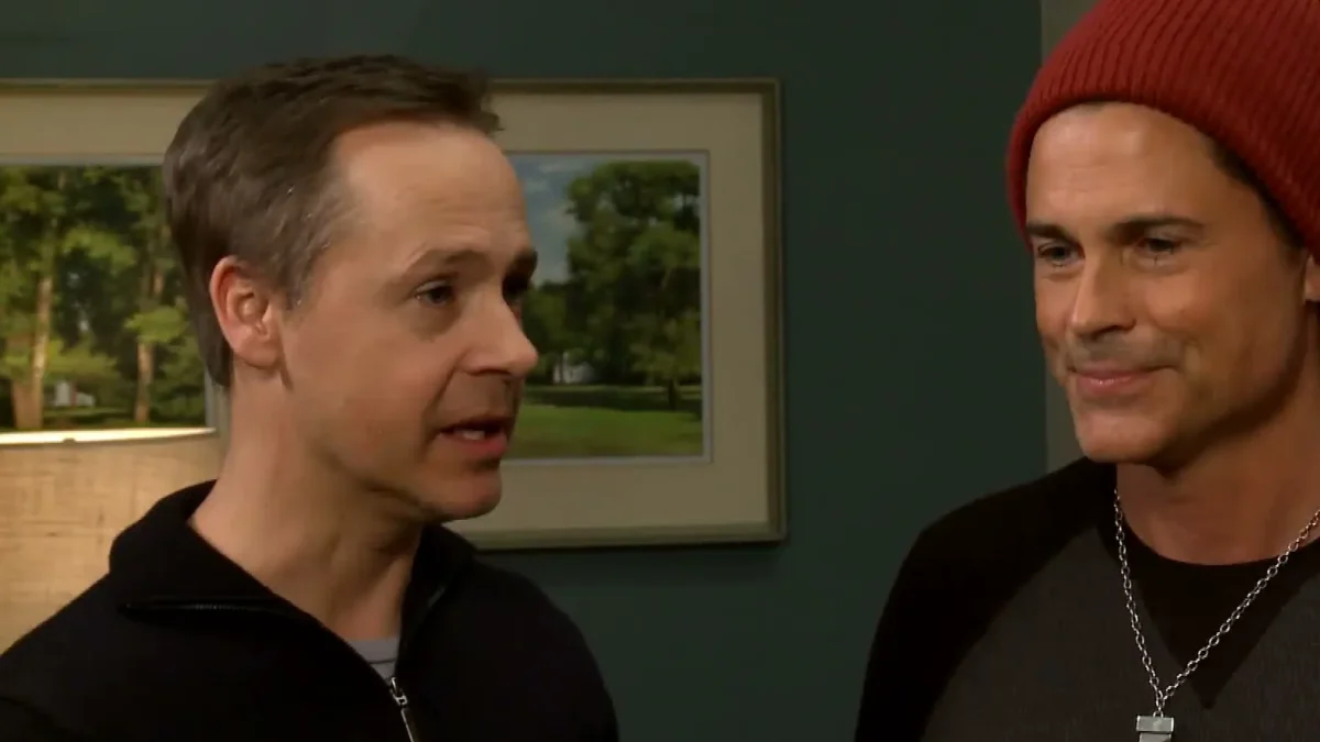 Chad Lowe in a black zip-up talking to his brother Rob Lowe, who wears a red knit cap, on a TV set