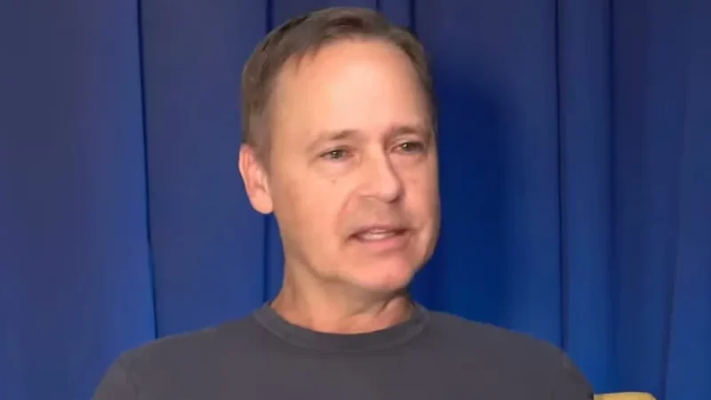 Chad Lowe in a gray T-shirt speaking in front of a blue curtain during a podcast interview