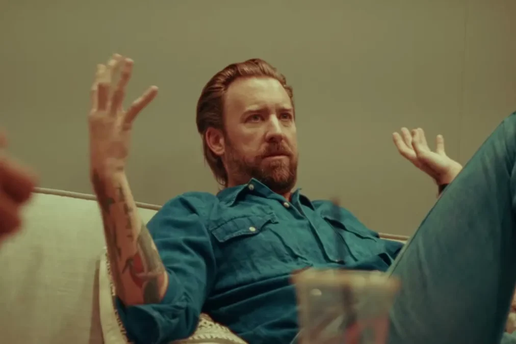 Charles Kelley in a teal denim shirt leaning back on a studio couch with his hands raised