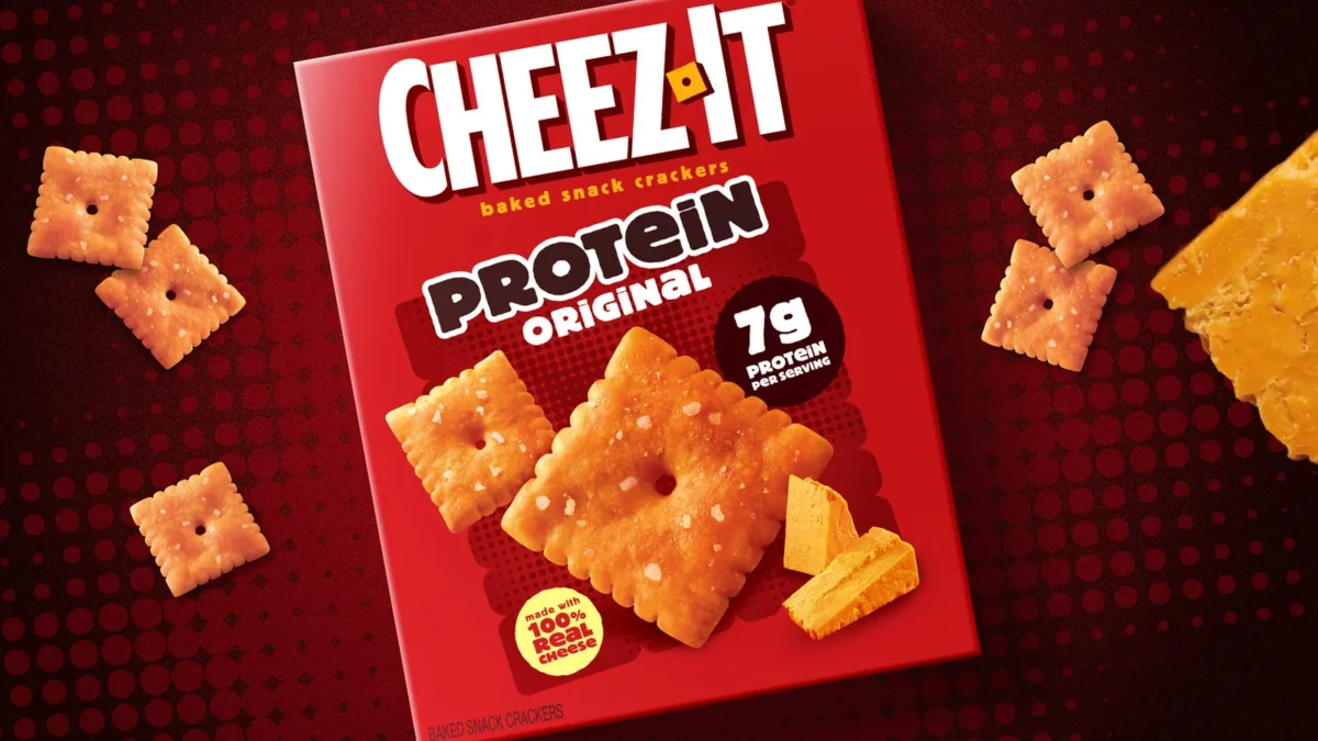 A red box of Cheez-It Protein Original baked snack crackers, with a 7g protein per serving badge and a Made with 100% real cheese seal, surrounded by Cheez-It crackers and a wedge of cheddar