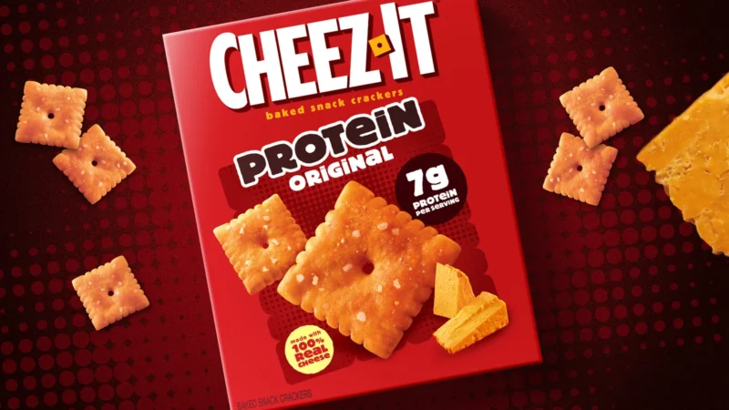 A red box of Cheez-It Protein Original baked snack crackers, with a 7g protein per serving badge and a Made with 100% real cheese seal, surrounded by Cheez-It crackers and a wedge of cheddar