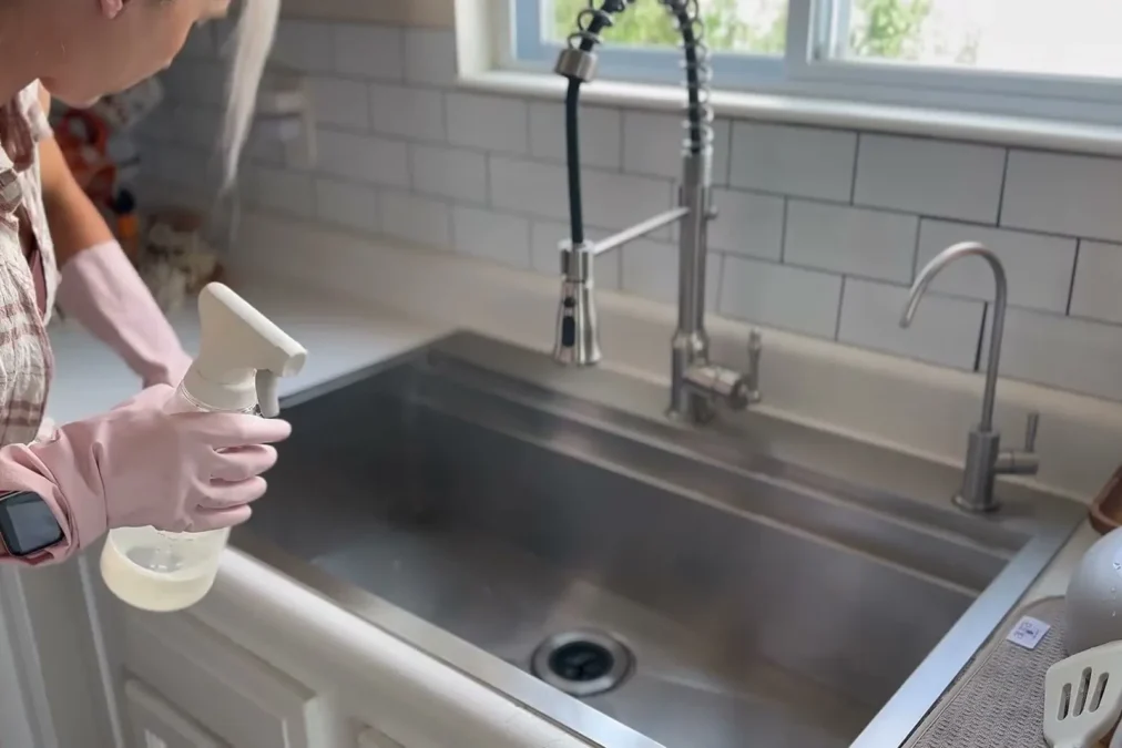 A person in pink rubber gloves spraying cleaner into a stainless steel kitchen sink