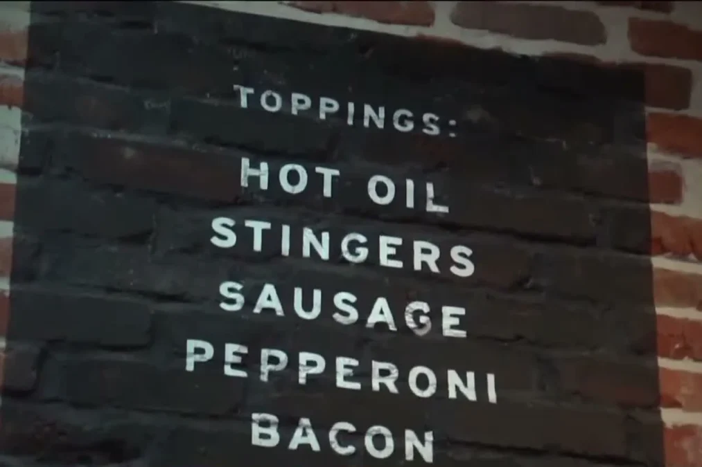 A painted toppings list on a brick wall at Colony Grill: hot oil, stingers, sausage, pepperoni, bacon