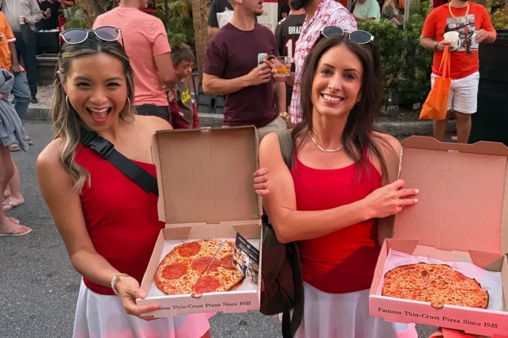 Two smiling women in red Buccaneers colors hold open boxes with a pepperoni pizza and a cheese pizza from Colony Grill at an outdoor event