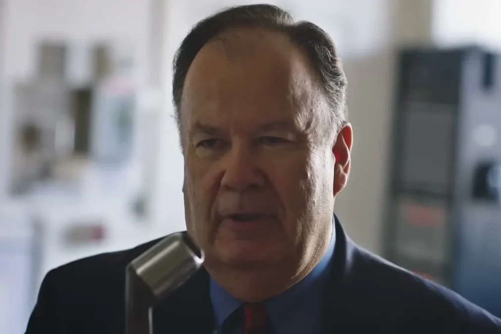 Dennis Haskins in a dark suit and red tie speaking into a vintage microphone in a school office, in Khalid's Young Dumb & Broke video