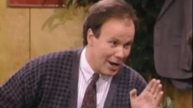 Dennis Haskins as Principal Richard Belding in a gray cardigan and tie, smiling and raising one hand in his office on Saved by the Bell