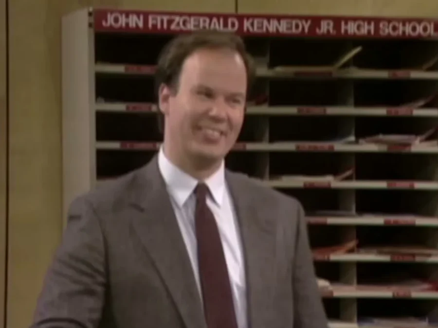 Dennis Haskins as Mr. Belding smiling in a brown suit in front of mail slots under a sign reading John Fitzgerald Kennedy Jr. High School
