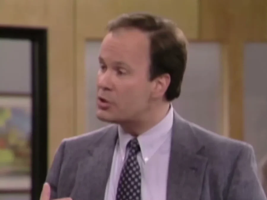 Dennis Haskins as Mr. Belding in a gray suit and polka-dot tie, talking in his office