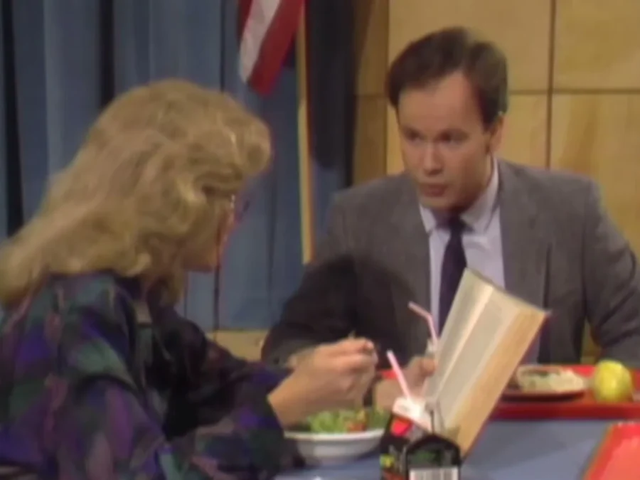 Dennis Haskins as Mr. Belding at a cafeteria table across from a woman with curly blond hair and glasses