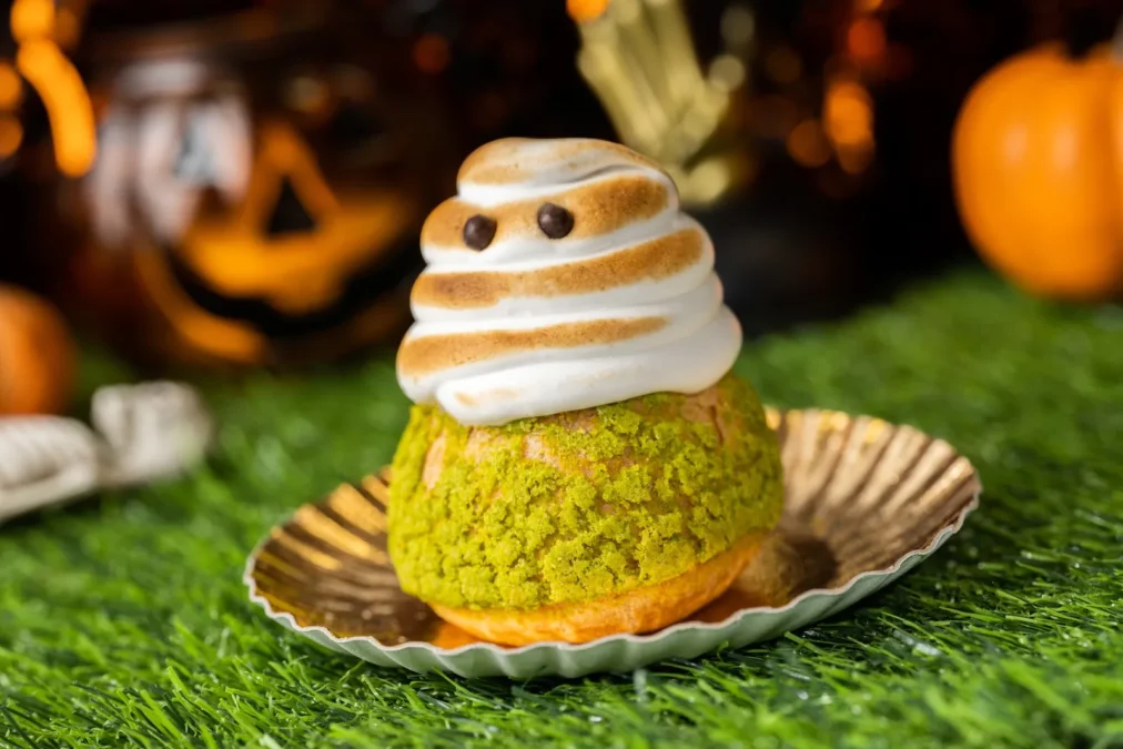The Ghost Cream Puff, a green-crusted cream puff topped with a toasted meringue ghost with chocolate eyes, on a gold paper liner