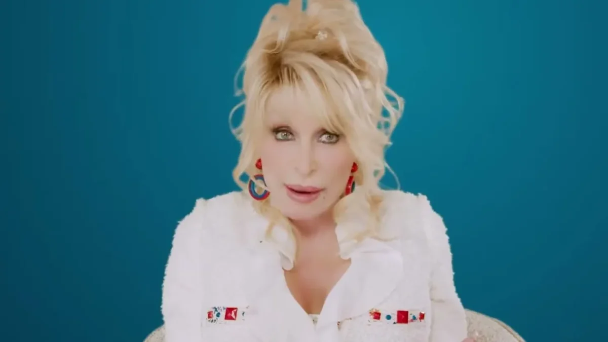 Dolly Parton in a white embroidered blouse speaking to the camera against a blue background