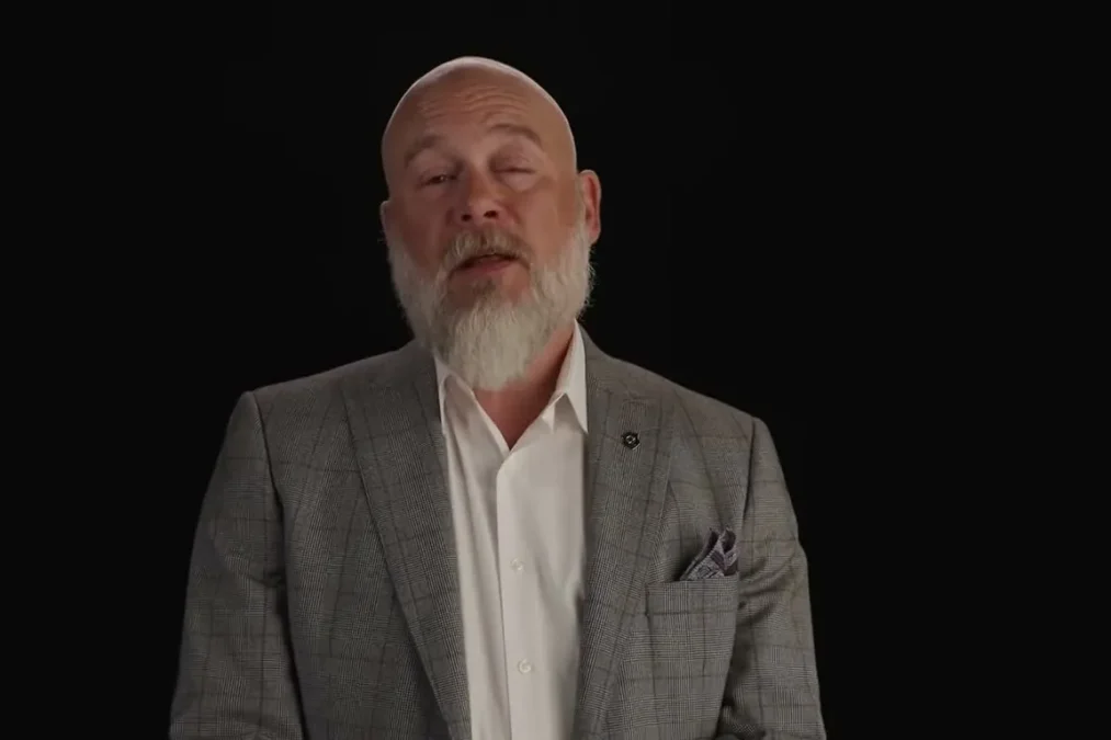 Bryan Seaver, bald with a white beard and a gray plaid jacket, speaking to the camera against a black background