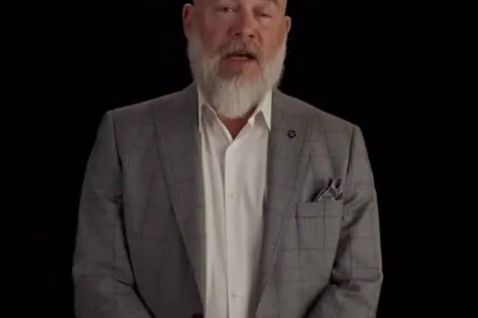 Bryan Seaver, bald with a white beard and a gray suit, speaking to the camera against a black background