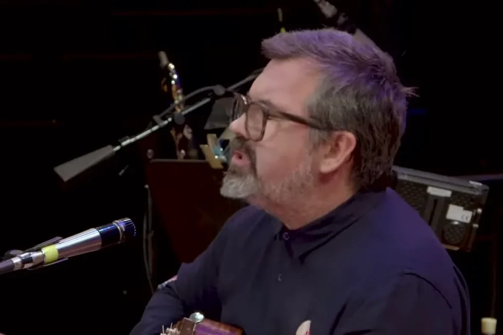 Duncan Sheik singing into a microphone while playing acoustic guitar