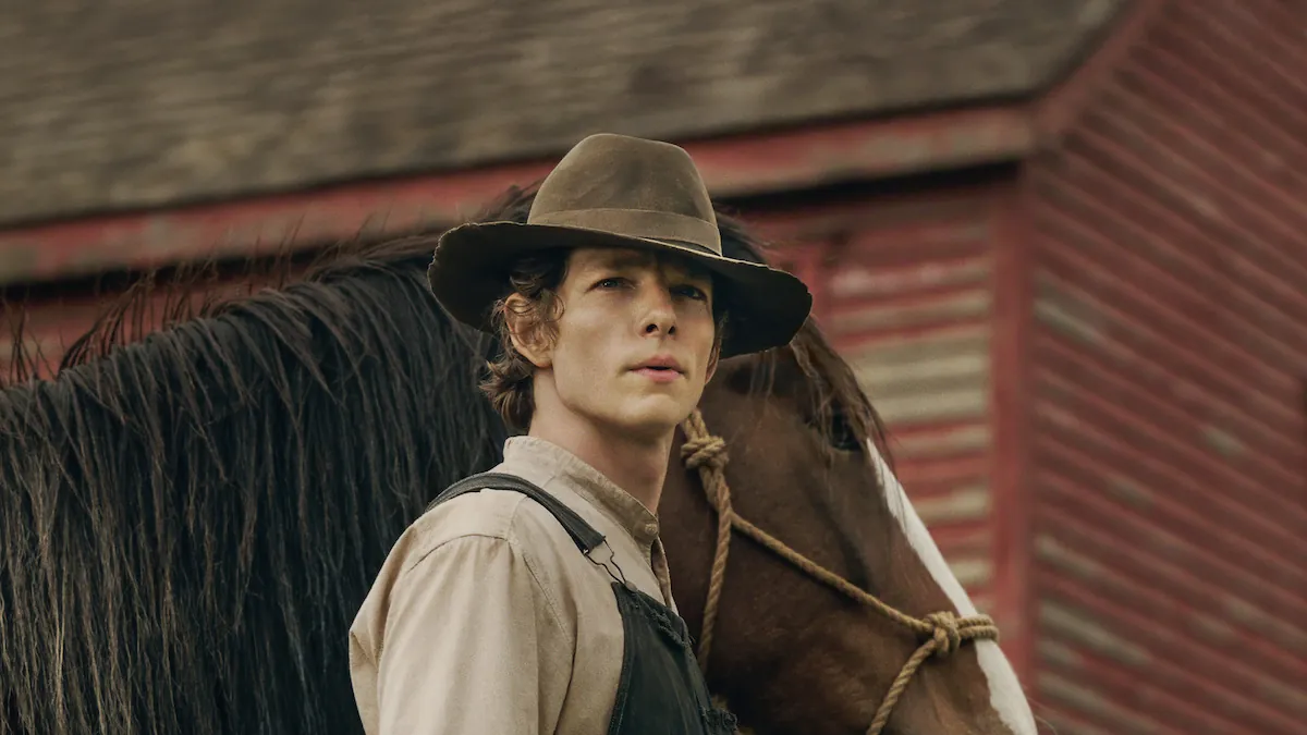 Mike Faist as Charles Trask in a hat and overalls holding a horse beside a red barn in East of Eden