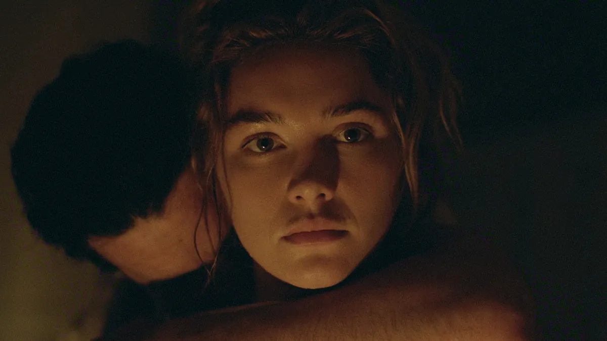 Florence Pugh as Cathy looking at the camera while a man embraces her in a dim room in East of Eden