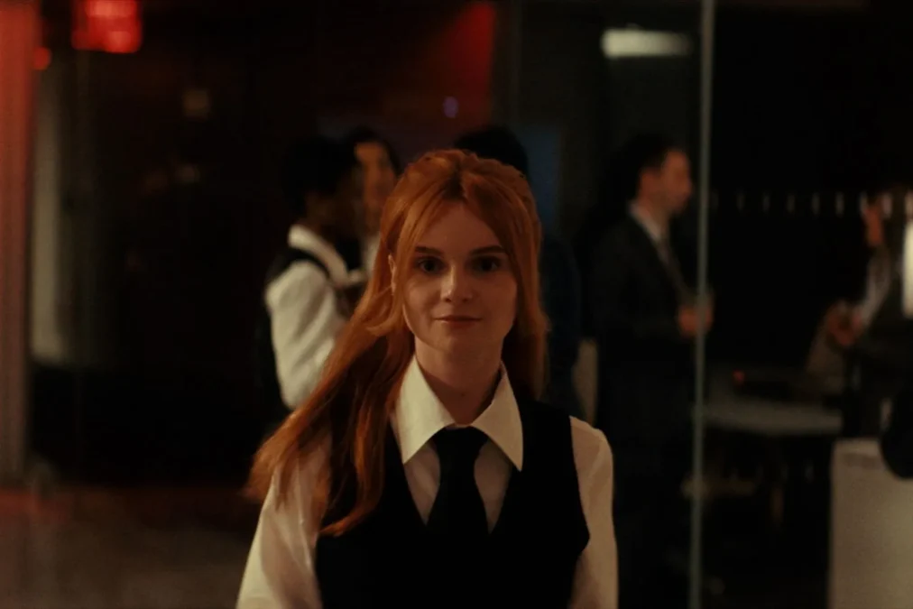 Lola Petticrew as Catherine, with long red hair and a black vest and tie, walks through a crowded lobby in Furious