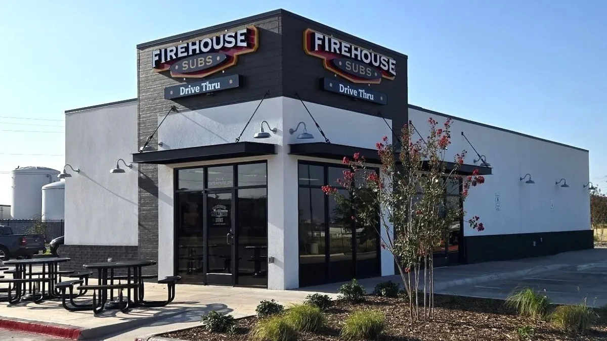 Exterior of a Firehouse Subs restaurant with a drive-thru, white walls, dark trim, red Firehouse Subs signs and outdoor picnic tables