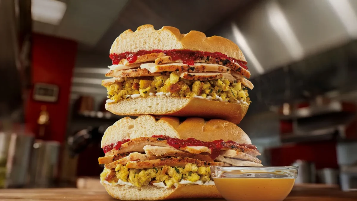 Two halves of a Firehouse Subs Thanksgiving Turkey Sub stacked on a wooden counter, with turkey, stuffing and cranberry sauce on a toasted roll and a cup of gravy beside them, in a restaurant kitchen