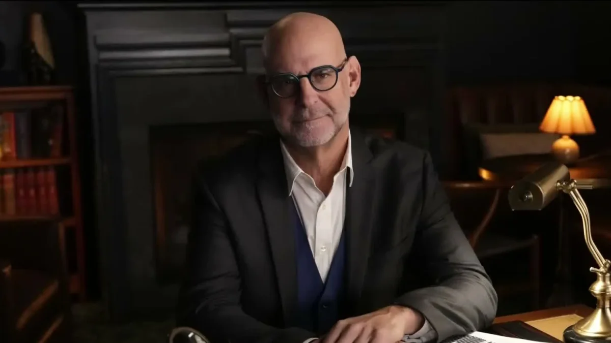 Harlan Coben in glasses and a dark jacket seated at a desk with a brass lamp in a wood-paneled study