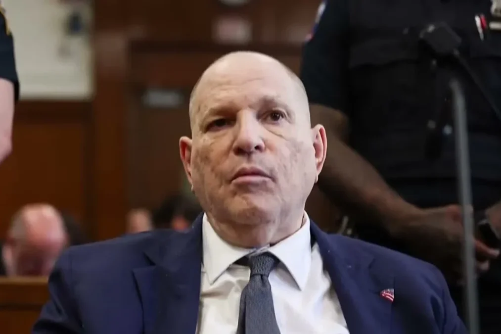 Harvey Weinstein, bald and in a navy suit, seated in court between court officers