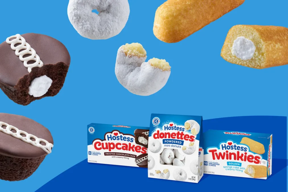 Hostess everyday snacks on a blue background: a bitten chocolate CupCake with its white Squiggle and creamy filling, a Twinkie cut open, powdered Donettes and boxes of CupCakes, Powdered Donettes and Golden Twinkies