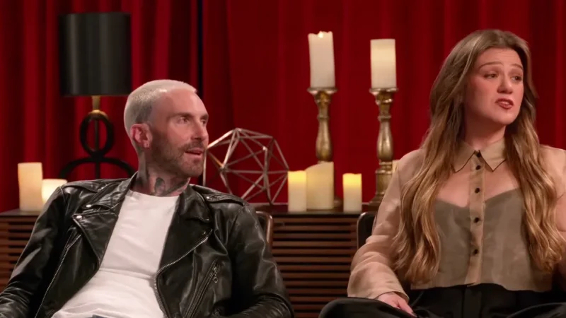Adam Levine in a black leather jacket and Kelly Clarkson in a sheer beige blouse, seated on a set with red curtains and candles