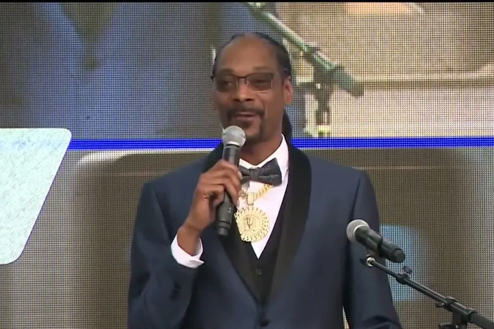 Snoop Dogg in a tuxedo and bow tie, wearing tinted glasses, speaking into a microphone at a podium