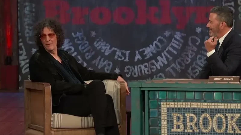 Howard Stern in tinted glasses sitting in a guest chair beside Jimmy Kimmel at a desk with a Brooklyn sign on Jimmy Kimmel Live