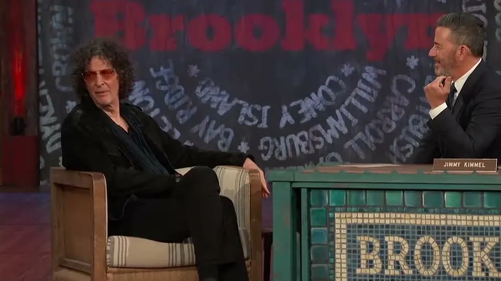 Howard Stern in tinted glasses sitting in a guest chair beside Jimmy Kimmel at a desk with a Brooklyn sign on Jimmy Kimmel Live