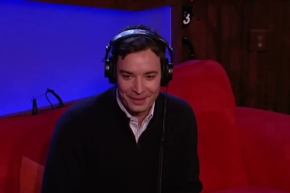 Jimmy Fallon in headphones smiling on a red couch in a radio studio