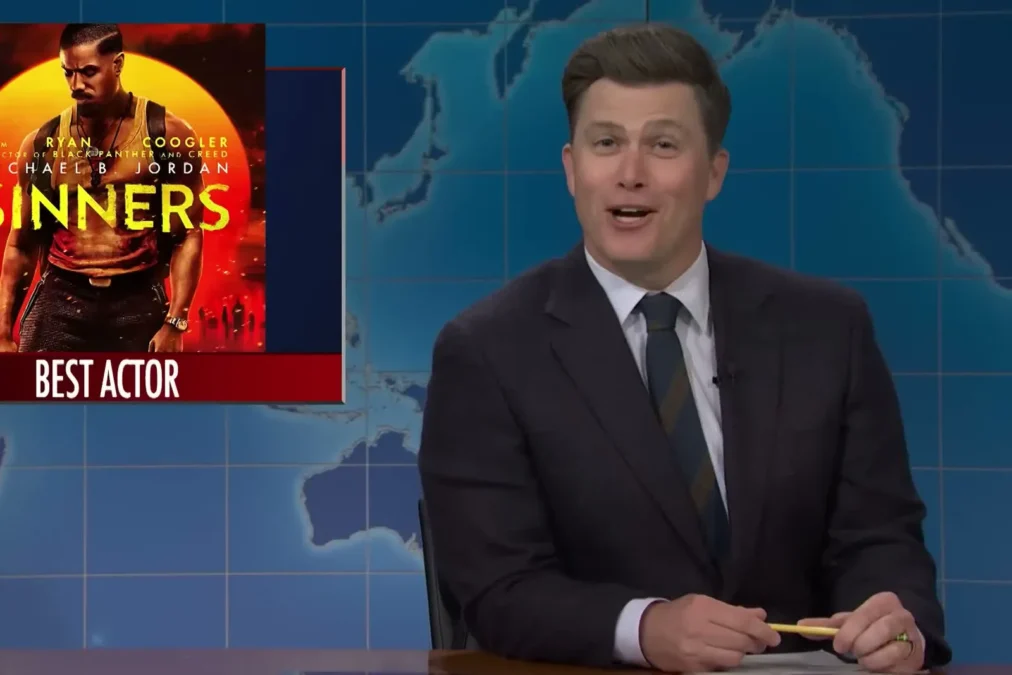 Colin Jost smiling at the Weekend Update desk on Saturday Night Live