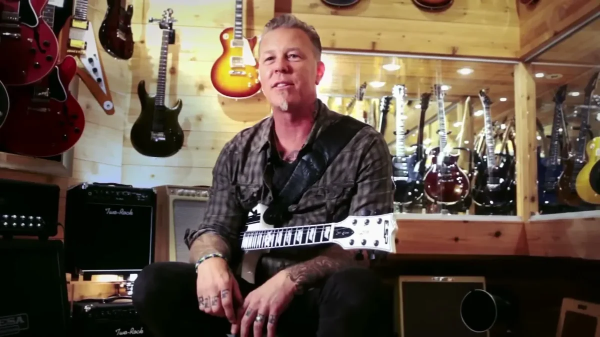 James Hetfield in a plaid shirt with a white guitar smiling in a guitar store