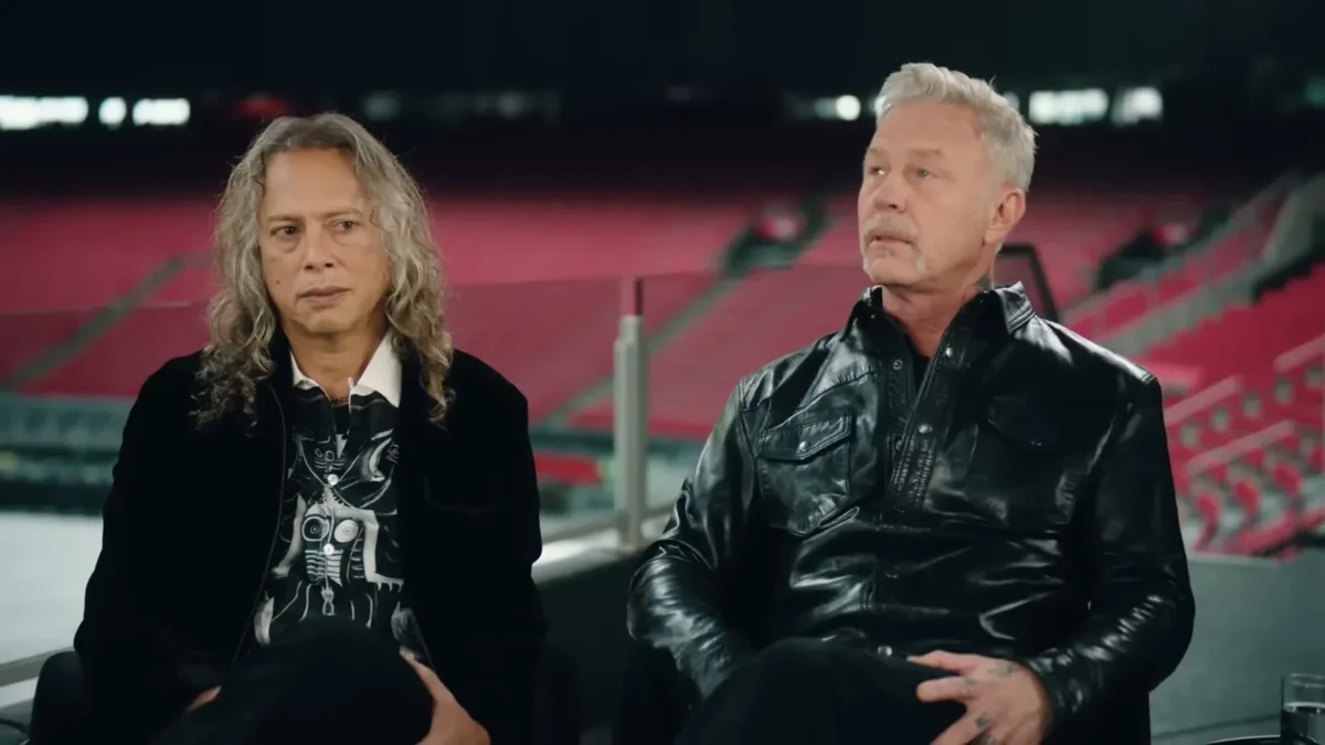 Kirk Hammett with long curly hair and James Hetfield in a leather jacket seated inside an empty stadium